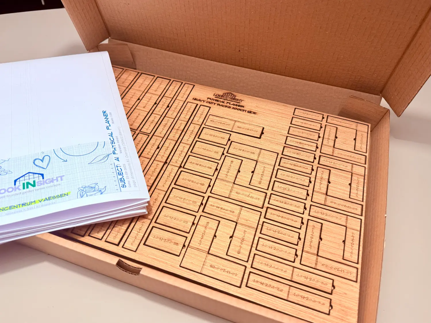 An opened cardboard mailer holding a laser-cut wooden sheet of heavy duty rack tiles, with the folded A1 plan sheet resting on top.