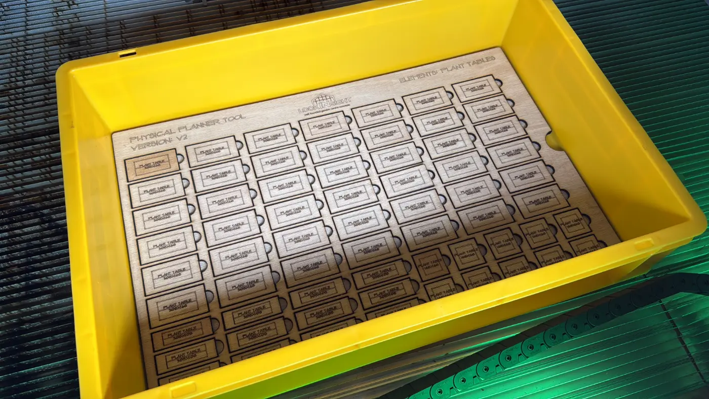An open yellow stacking crate containing a laser-cut sheet of plant table tiles, labelled Physical Planner Tool version V2.
