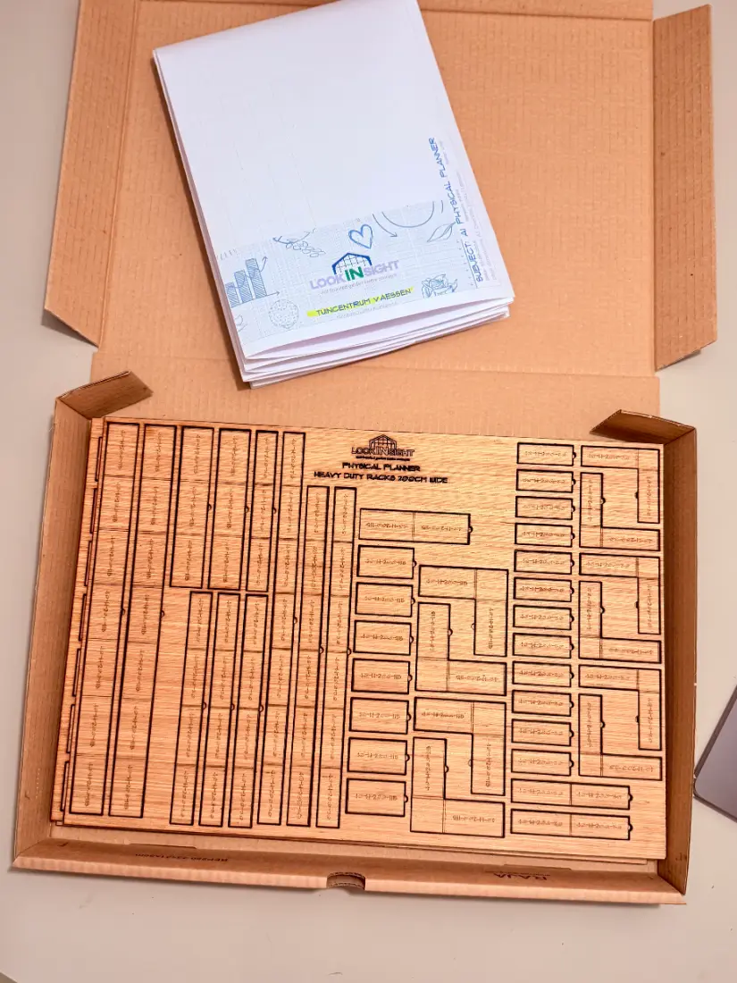 Top-down view of the opened mailer box with the wooden tile sheet inside and the A1 plan booklet beside it.