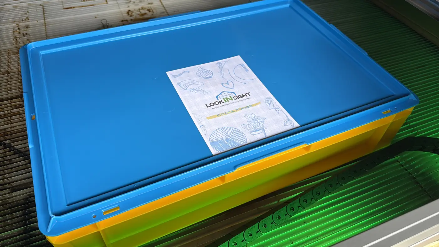 A closed blue-lidded stacking crate with a printed lookINsight Physical Planner label, sitting on the bed of a laser cutter.