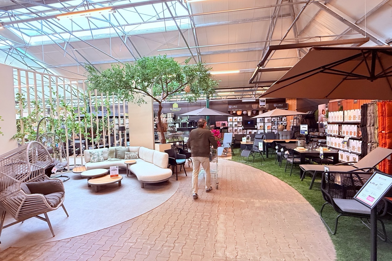 Tuincentrum De Bosrand: a customer walks the curved main route through the garden furniture department, past a lounge zone on one side and a dining zone on artificial grass on the other